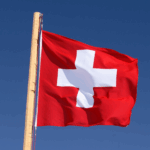 Swiss national flag in front of blue skies on wooden pole.