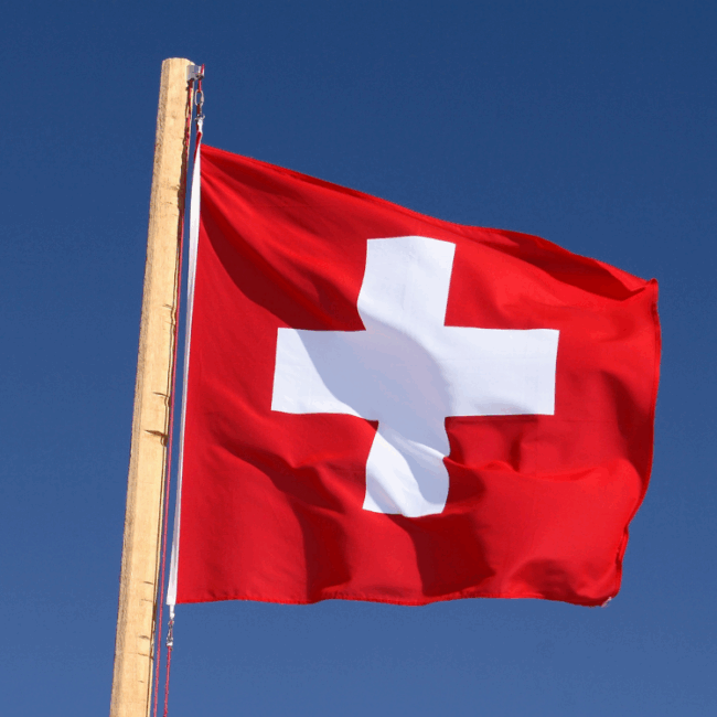 Swiss national flag in front of blue skies on wooden pole.